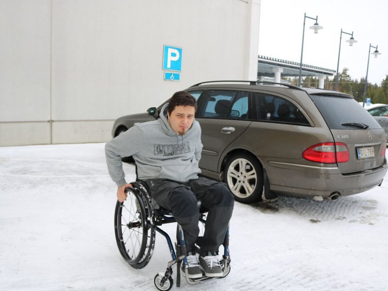 Mies pyörätuolissa auto parkkeerattu invapaikalle.